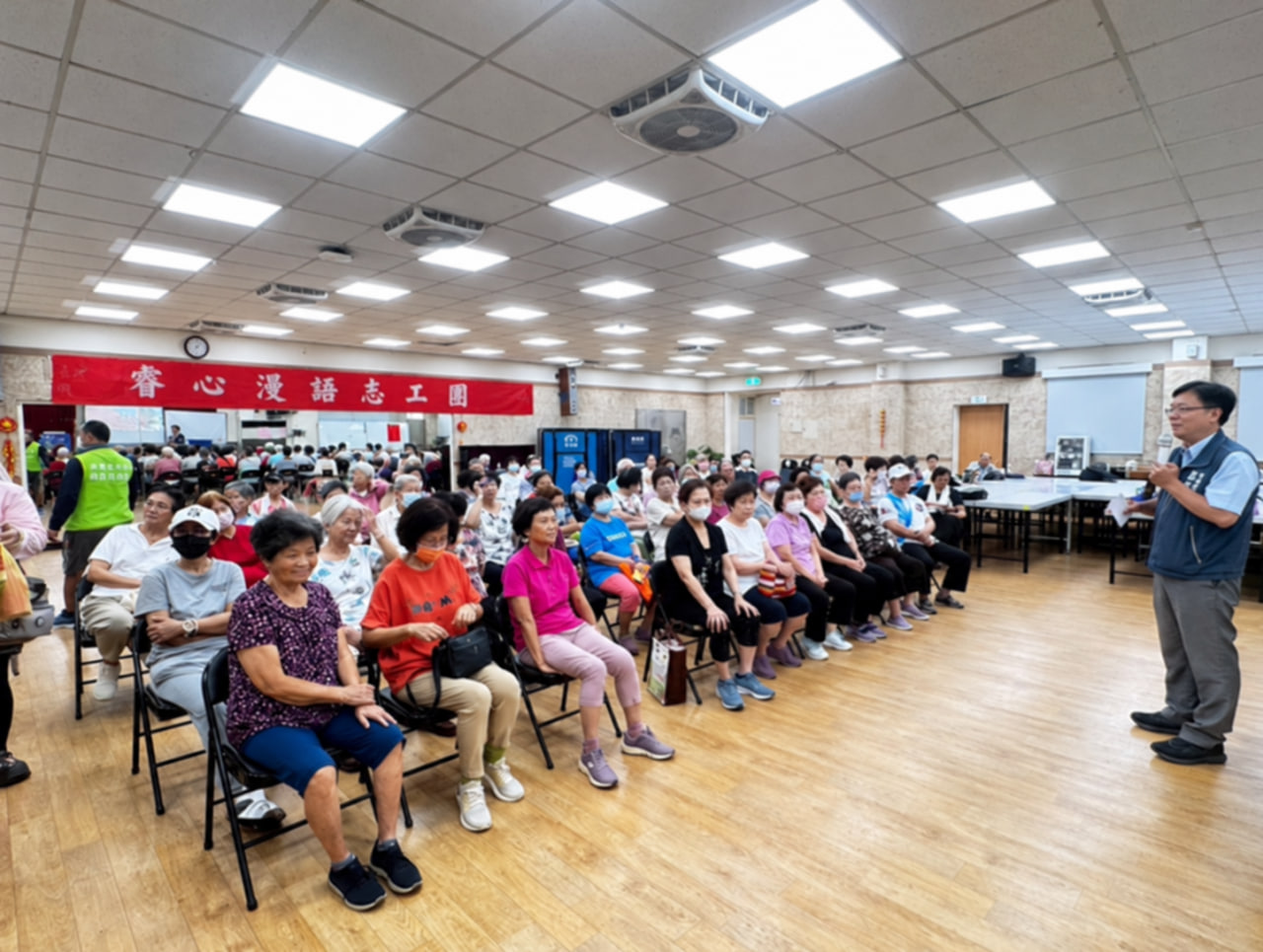 活動現場，參與者坐在室內聆聽講者分享，後方掛有志工團布條。
