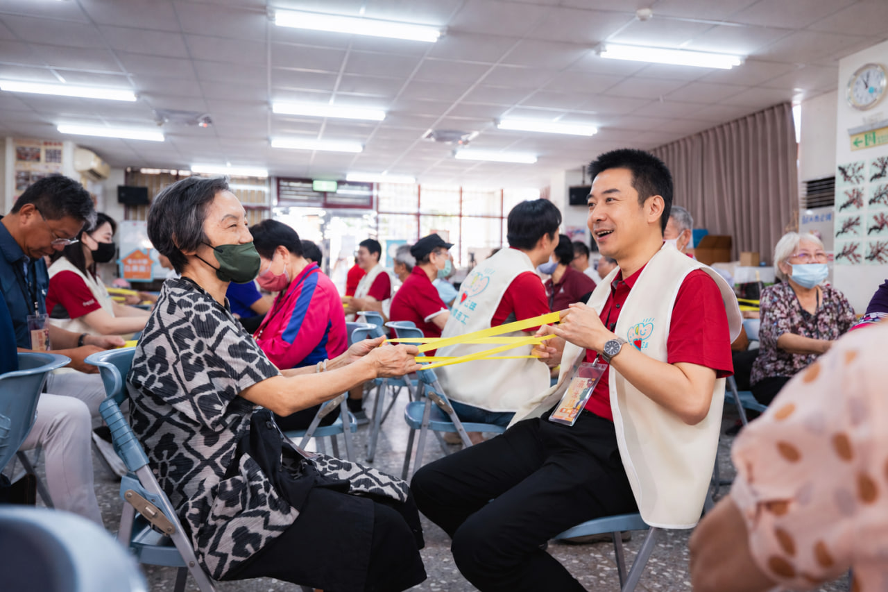 志工與長者面對面坐著，使用黃色彈力帶互動，周圍也有其他志工與長者參與活動。